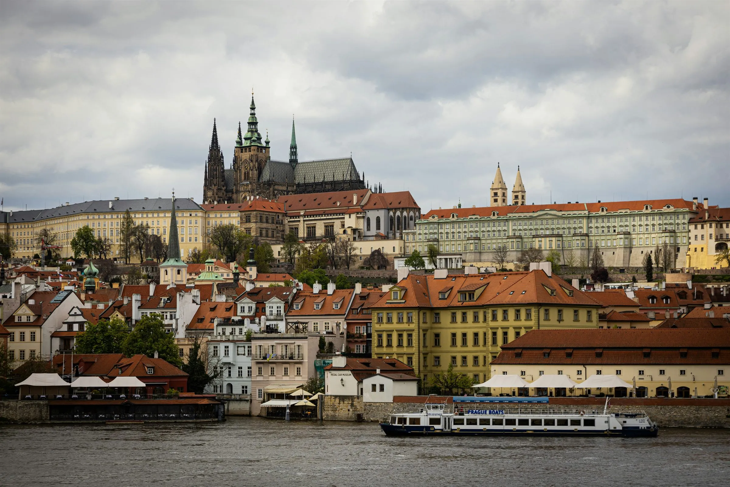 Prague Castle and St. Vitus Cathedral above the red roofs of Malá Strana, seen across the Vltava under a grey, cloudy sky.